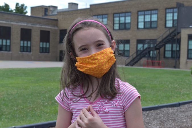 A girls poses in a face mask outside an elementary school.